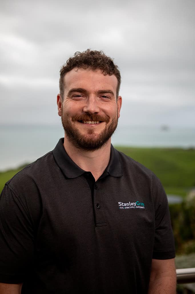 Stanley Gray civil engineer outdoors, smiling, with coastal landscape in background, wearing branded polo shirt.