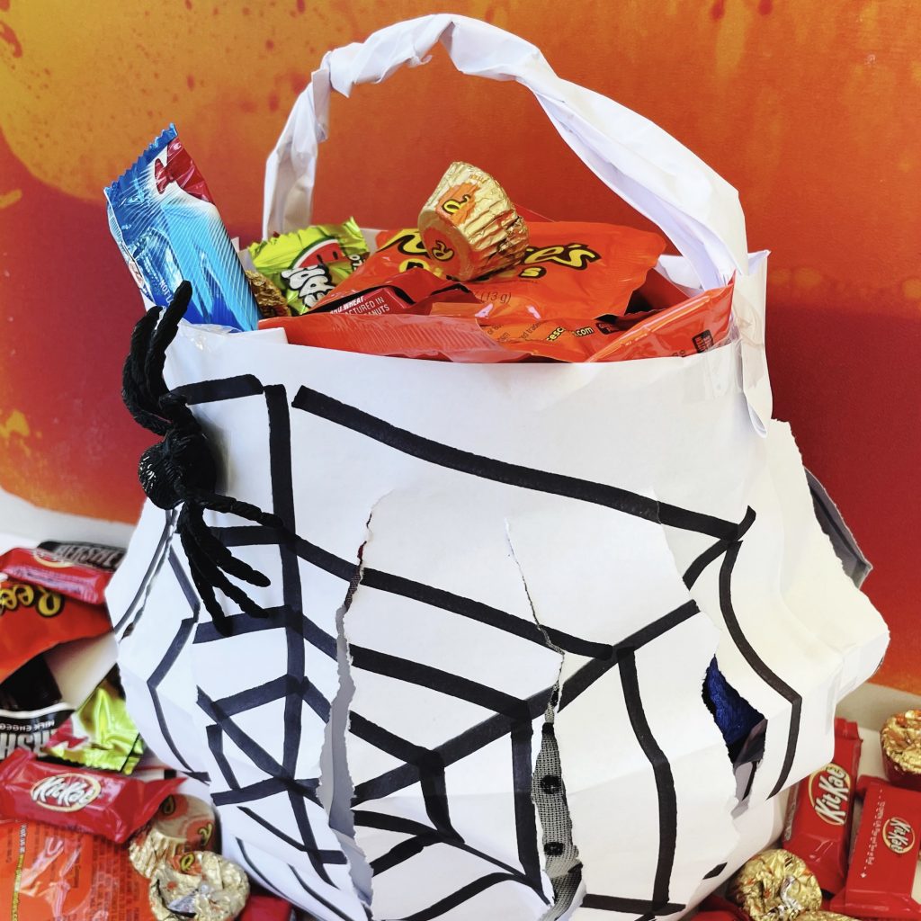 This image shows a Halloween candy bucket constructed from copy paper and tape. It's full of candy with an orange background.