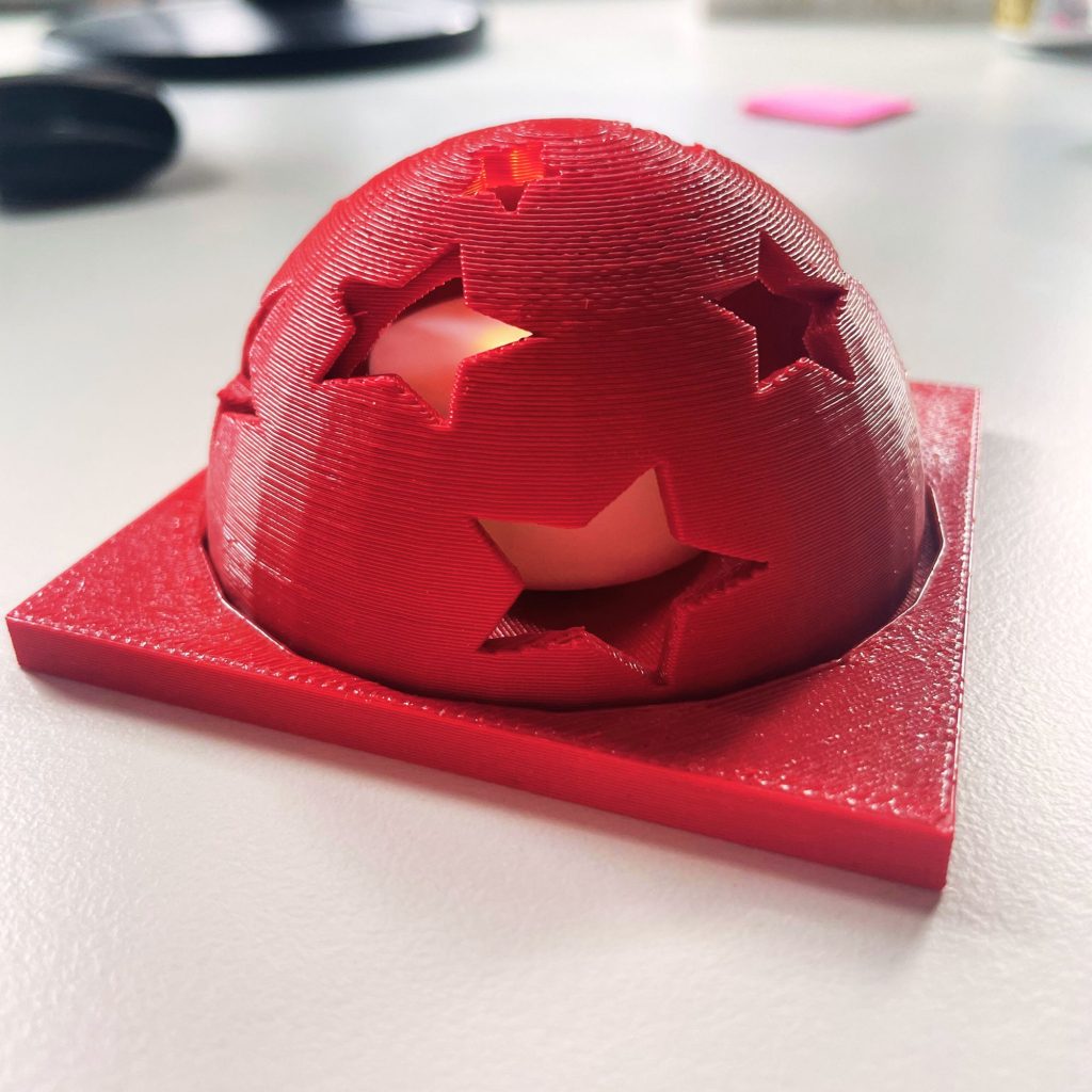 This image shows a red 3D printed lantern. There is a square base with a dome on top. The dome has star-shaped holes. Light is coming through from a tea light placed inside the dome.