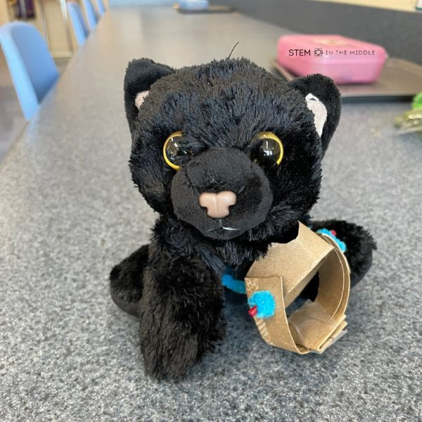 This image shows a black stuffed cat missing a front limb. It has a DIY prosthesis attached made from cardboard, pipe cleaners, and straws.