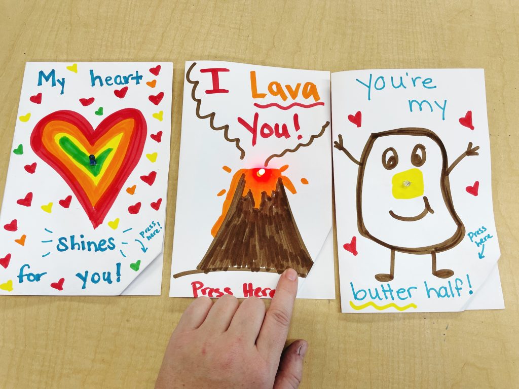 This image shows 3 DIY Valentines Day Cards with LED lights on the front. There is a hand pushing a button on the center card to light the LED.