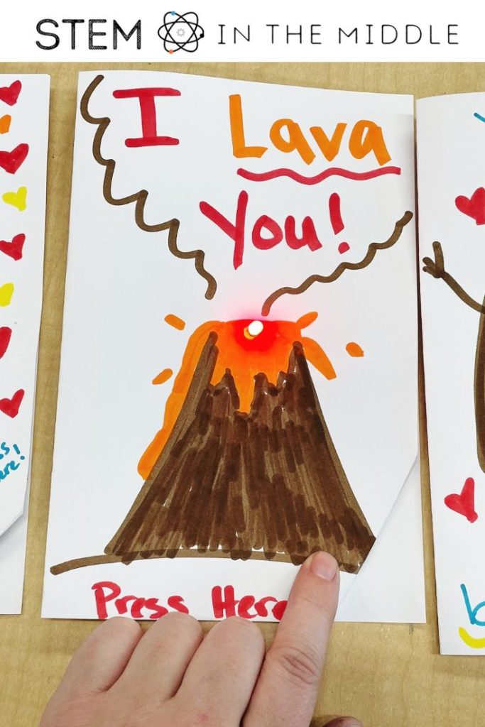 The image shows a DIY Valentine's Day card with a volcano and text that reads "I lava you!" A finger is pressing a button on the card that lights up a mini LED bulb on the volcano.