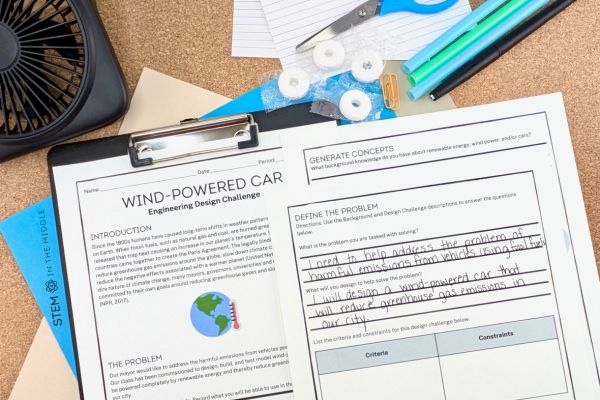 This image shows worksheets titled "Wind Powered Car: Engineering Design Challenge." The worksheets are partially completed. Surrounding the worksheets are a fan, scissors, pens, index cars, and life savers.