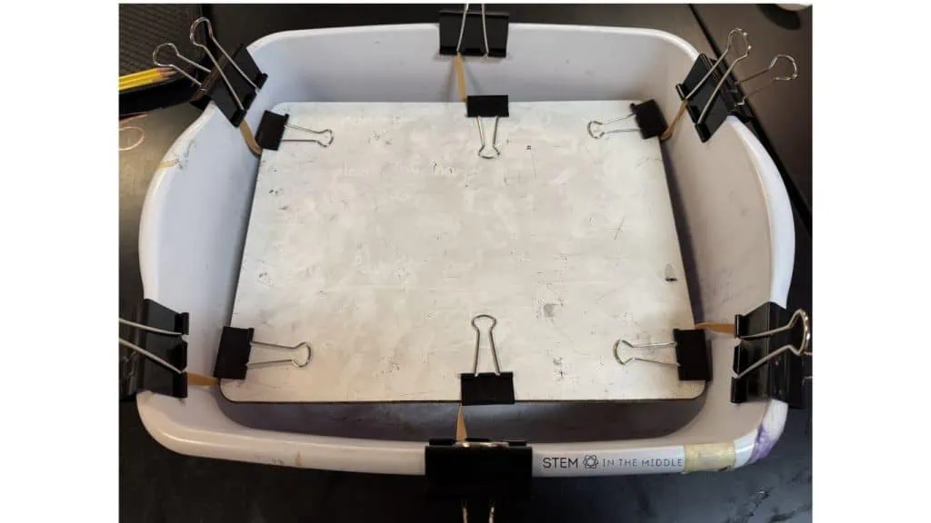 The image shows a DIY shake table created by suspending a whiteboard inside a larger box using rubber bands and binder clips.