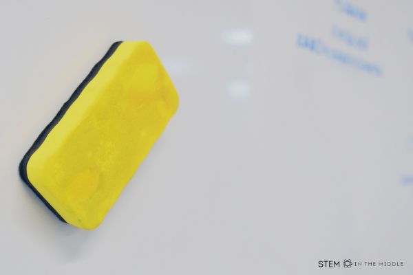 This image shows a yellow white board eraser sticking to a white board. There is some blue writing out of focus on the right side of the board.