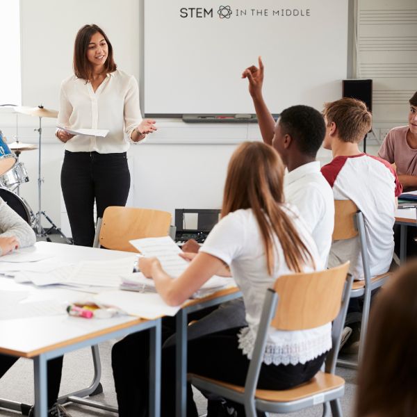 This image shows a teacher at the front of the classroom with teen students facing the teacher. One student has their hand raised.