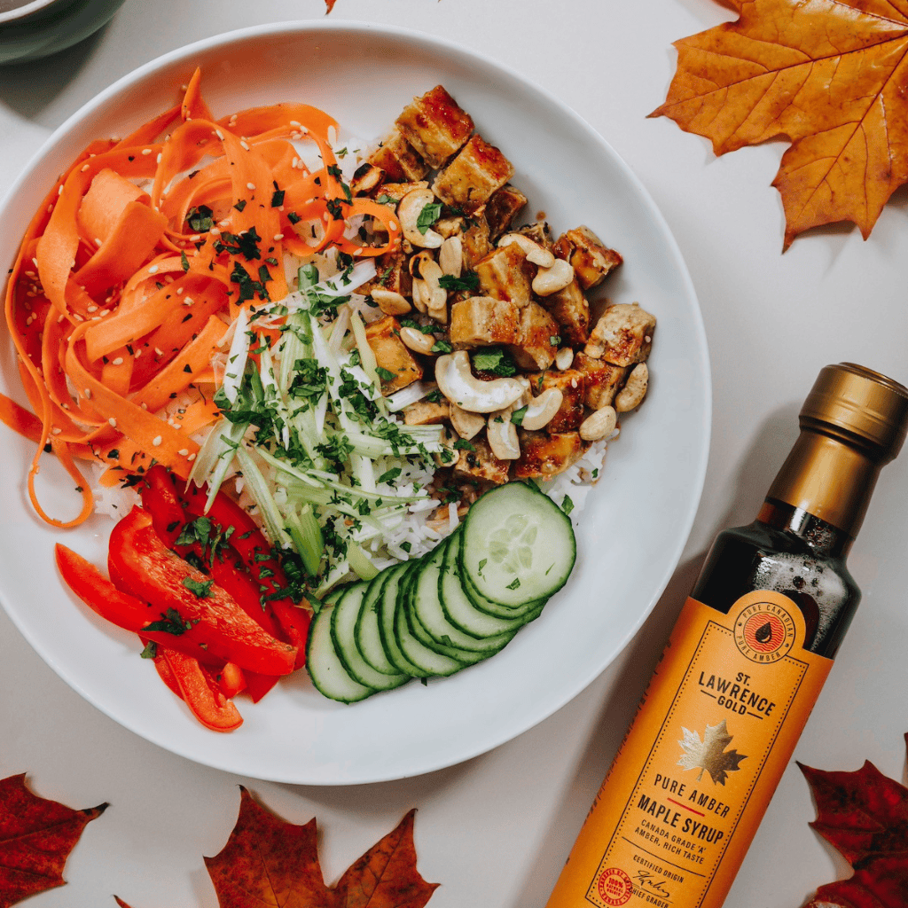 Tofu bowl with fresh vegetables and St Lawrence Gold Amber maple syrup next to it. Scattered maple leaves around.