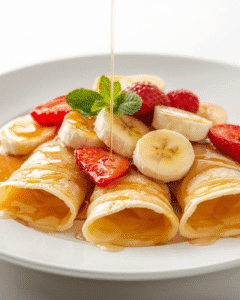 Maple syrup drizzling over crepe pancakes topped with strawberries and bananas