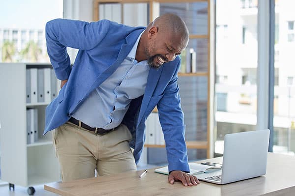 Experiencing back pain at work, man holding lower back in office environment, highlighting stress and pain relief treatment options.