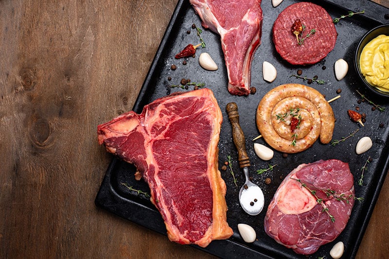 Succulent raw steaks and sausage on black stone serving platter, fresh herbs and garlic for healthy eating, New Wave Stress and Pain Relief, quality meat cuts for wellness and relaxation.
