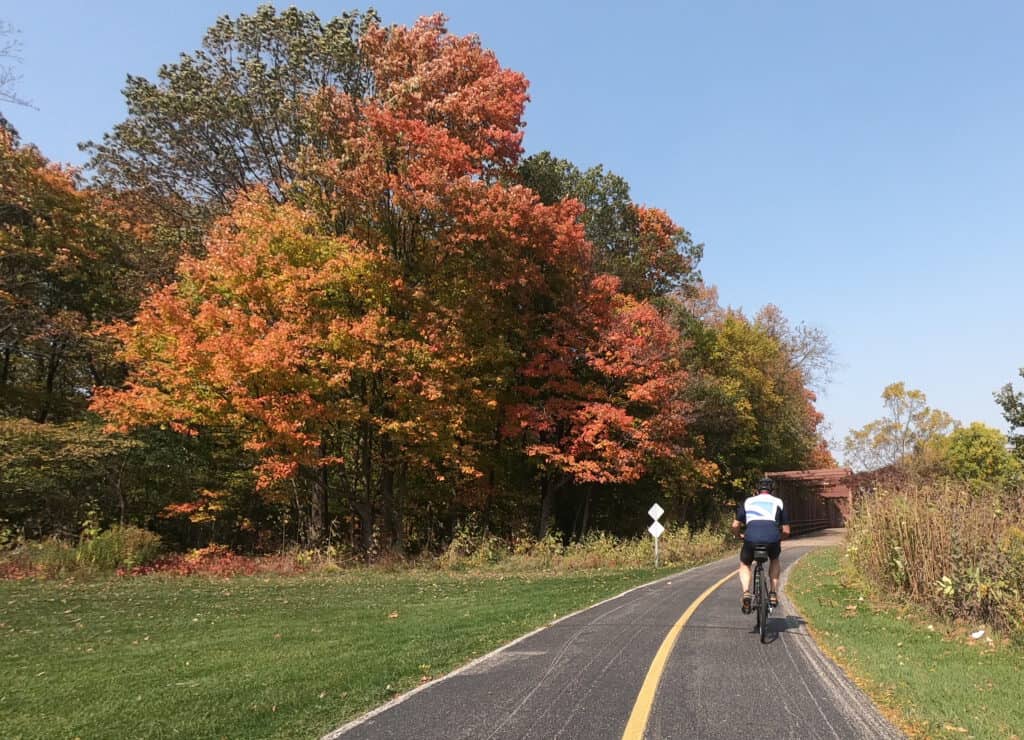 A bicyclist in shorts and jersey rides on a trail approaching colorful fall trees and a bridge in the distance.