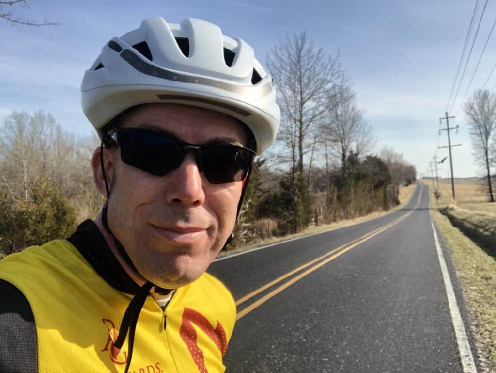 A man up close in a bicycle helmet and sunglasses with a yellow jersey stands in front of an open country road.