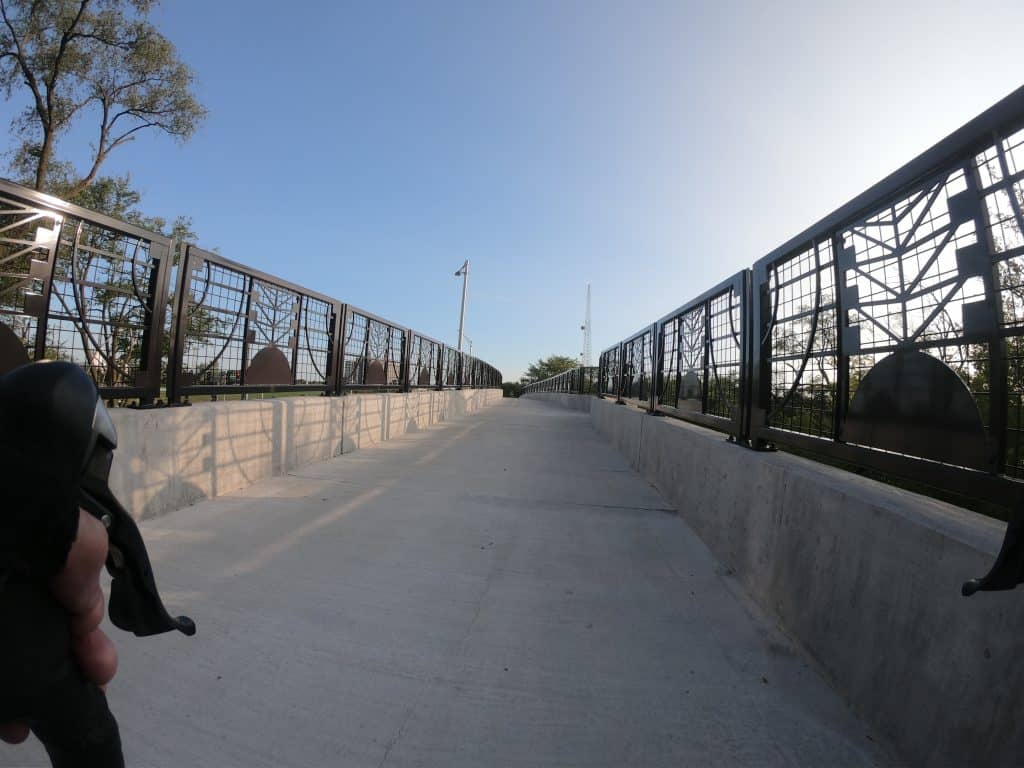 Looking forward at a cement path on a bridge with metal decorative railings and a sunny blue sky in the background.