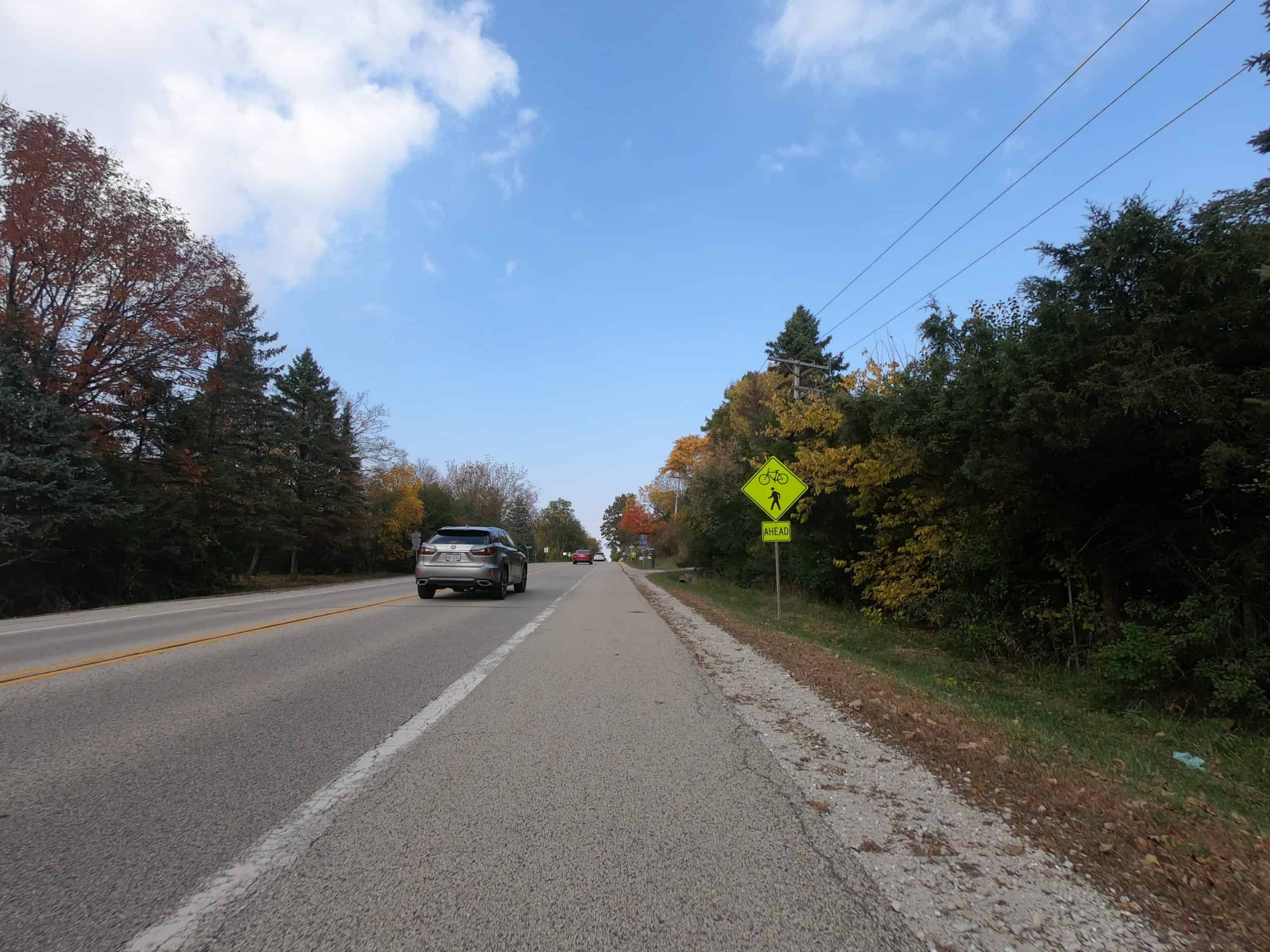 Looking down a colorful tree lined two-lane road with a car and yellow sign.