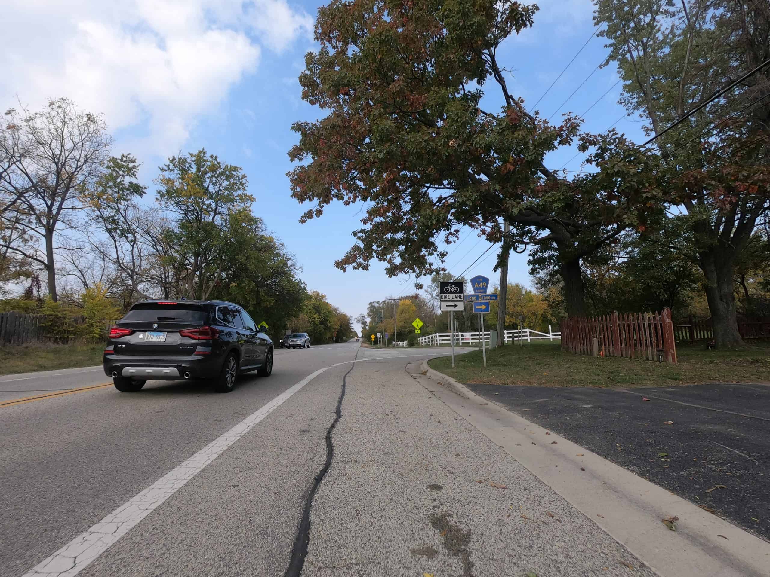 Looking down the shoulder of a colorful tree lined two-lane road with a car on our left.
