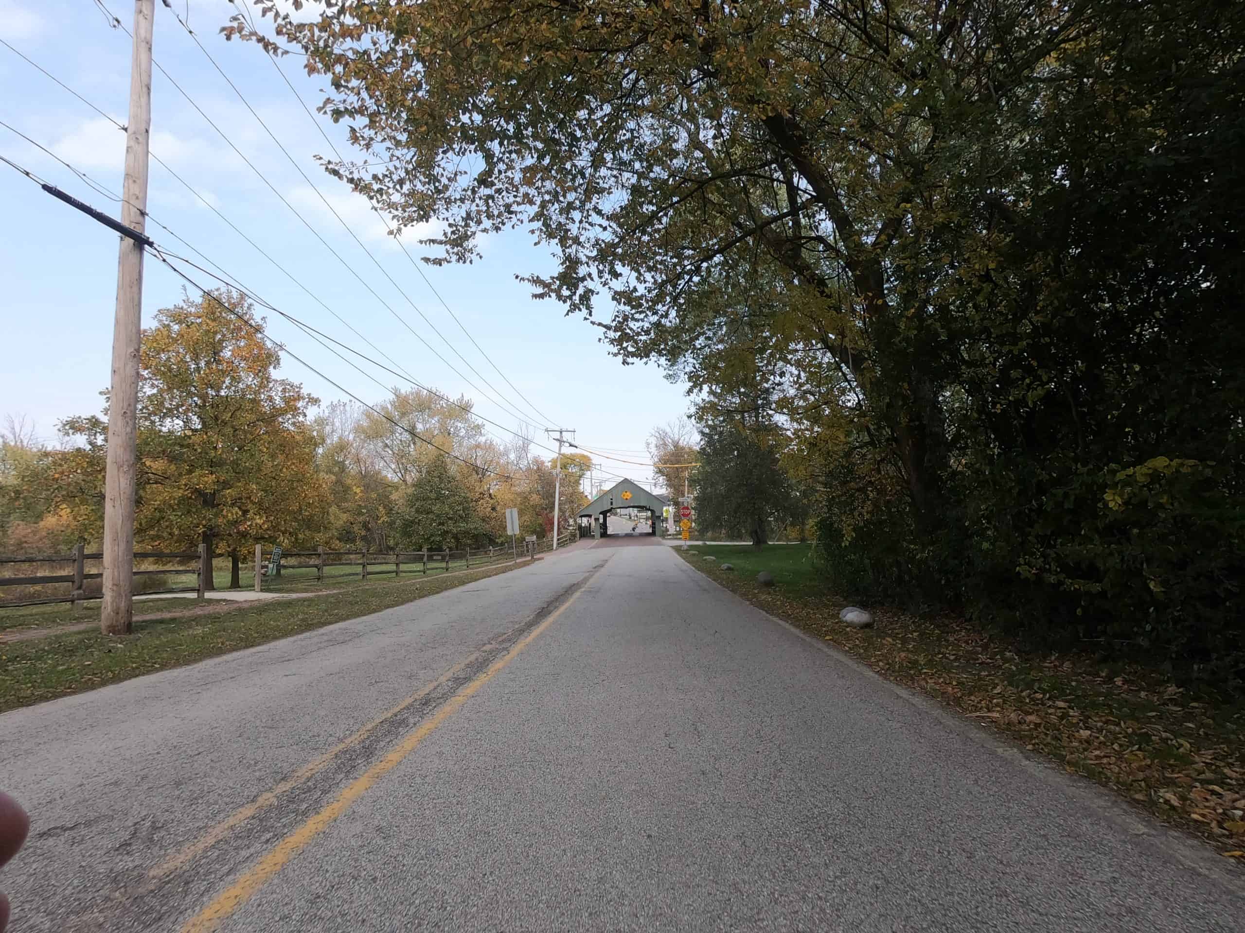 Looking down a colorful tree lined two-lane road with a covered bridge in the distance.