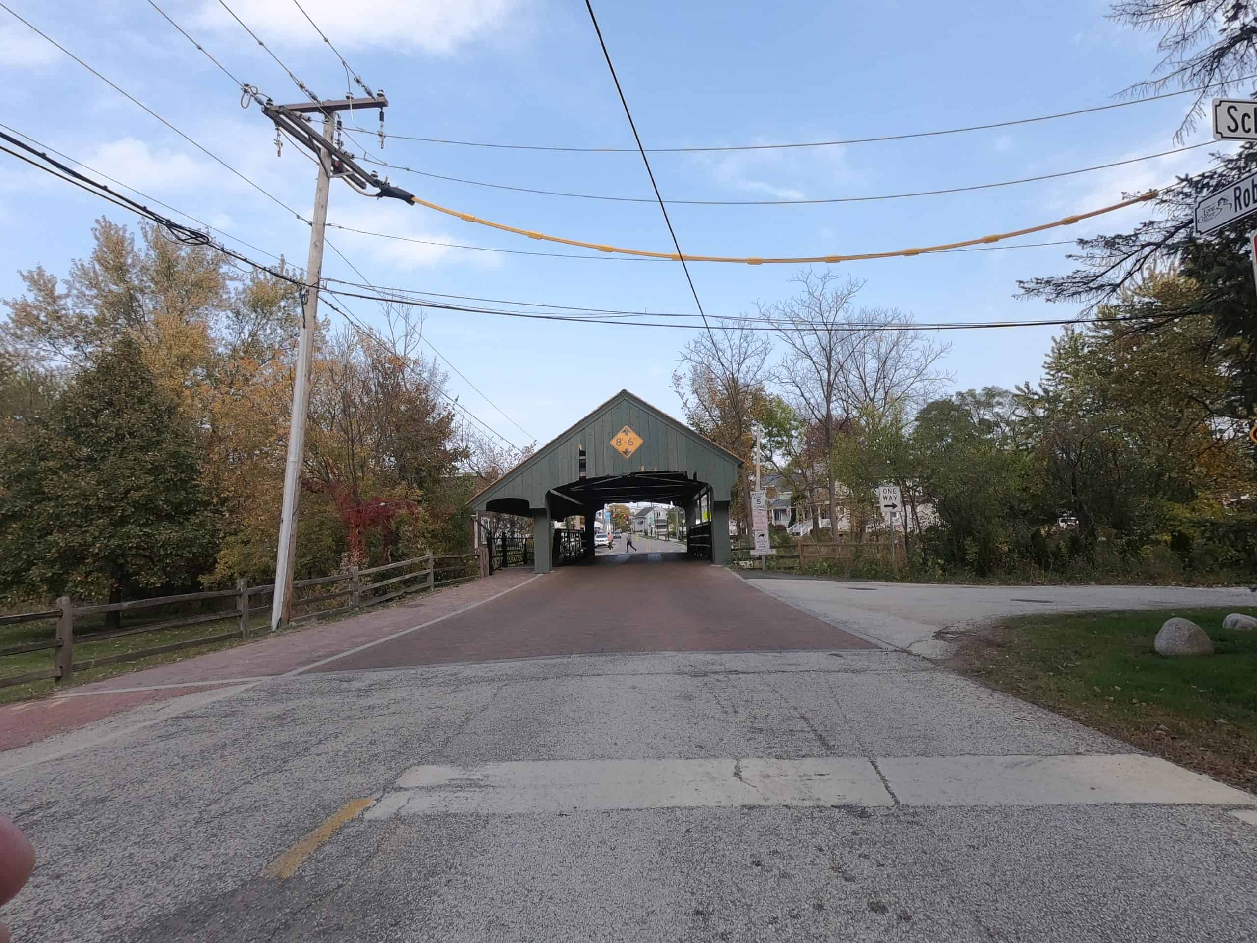 Looking down a street coming upon brick pavement and a gray covered bridge.