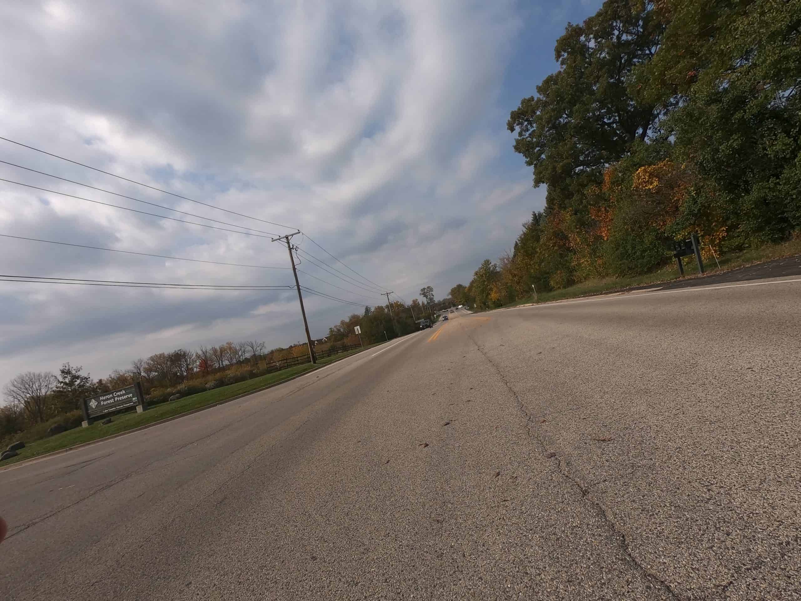 Looking down a colorful tree and meadow lined two-lane road on a tilt, coming on a forest preserve entrance.