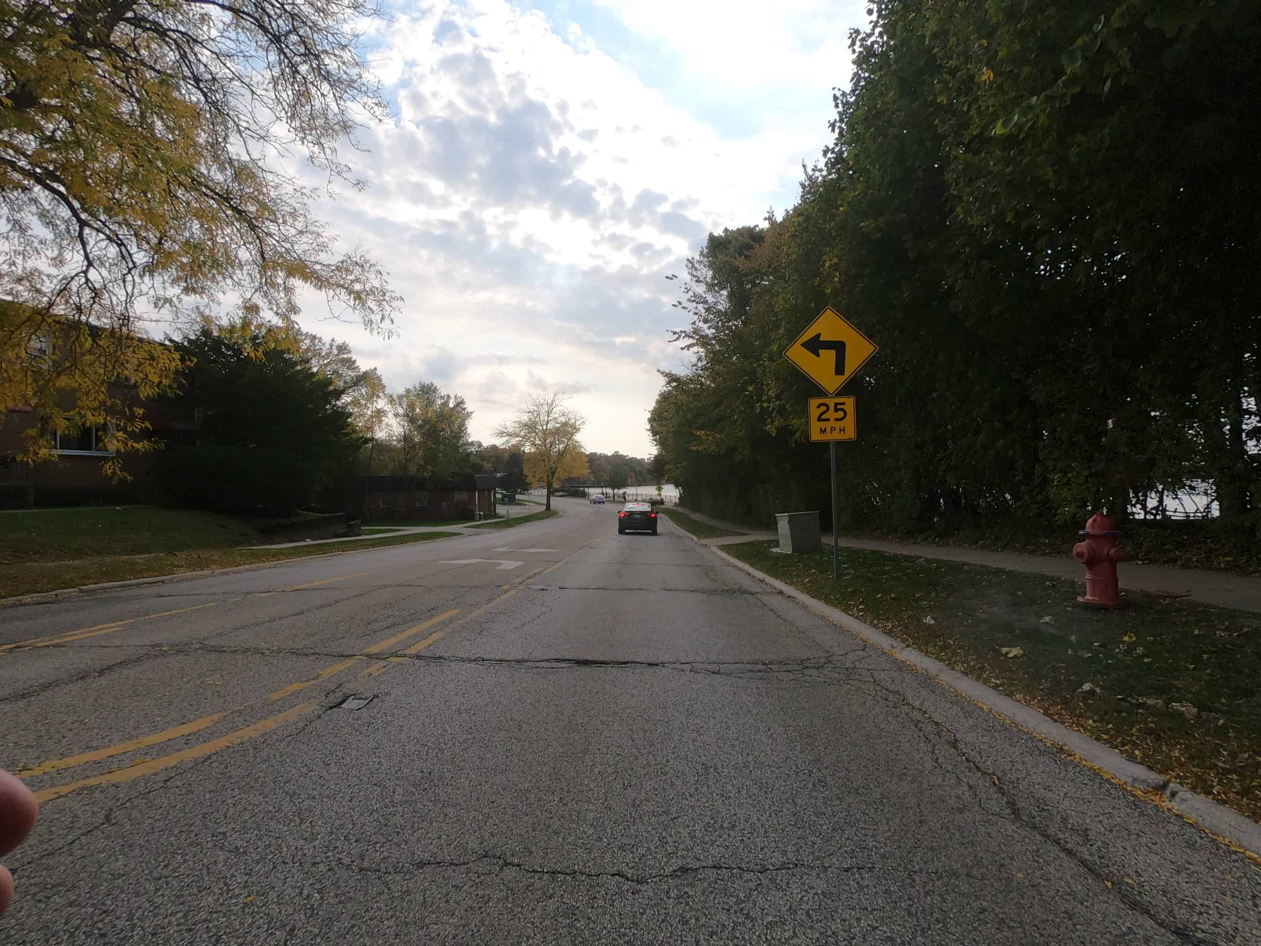 Looking down a cracked street with a lake on the right and some white clouds in front of the sun and blue sky.