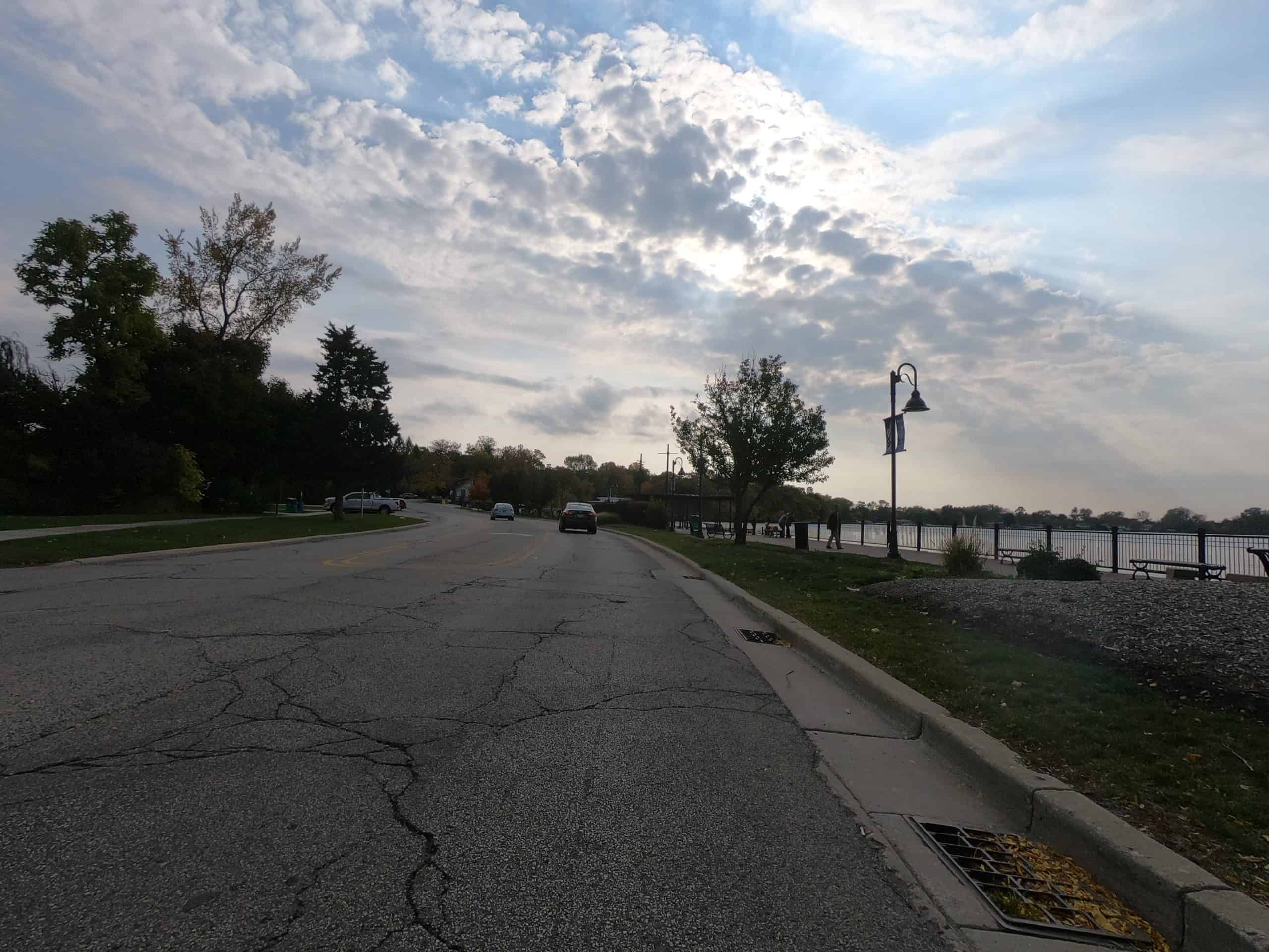 Looking down a cracked street with a lake on the right and some white clouds in front of the sun and blue sky.