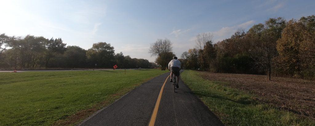 A bicyclist in a sweatshirt and casual shorts rides down a paved trail with a yellow line alongside lawn and a street on the left with a forest in the background.