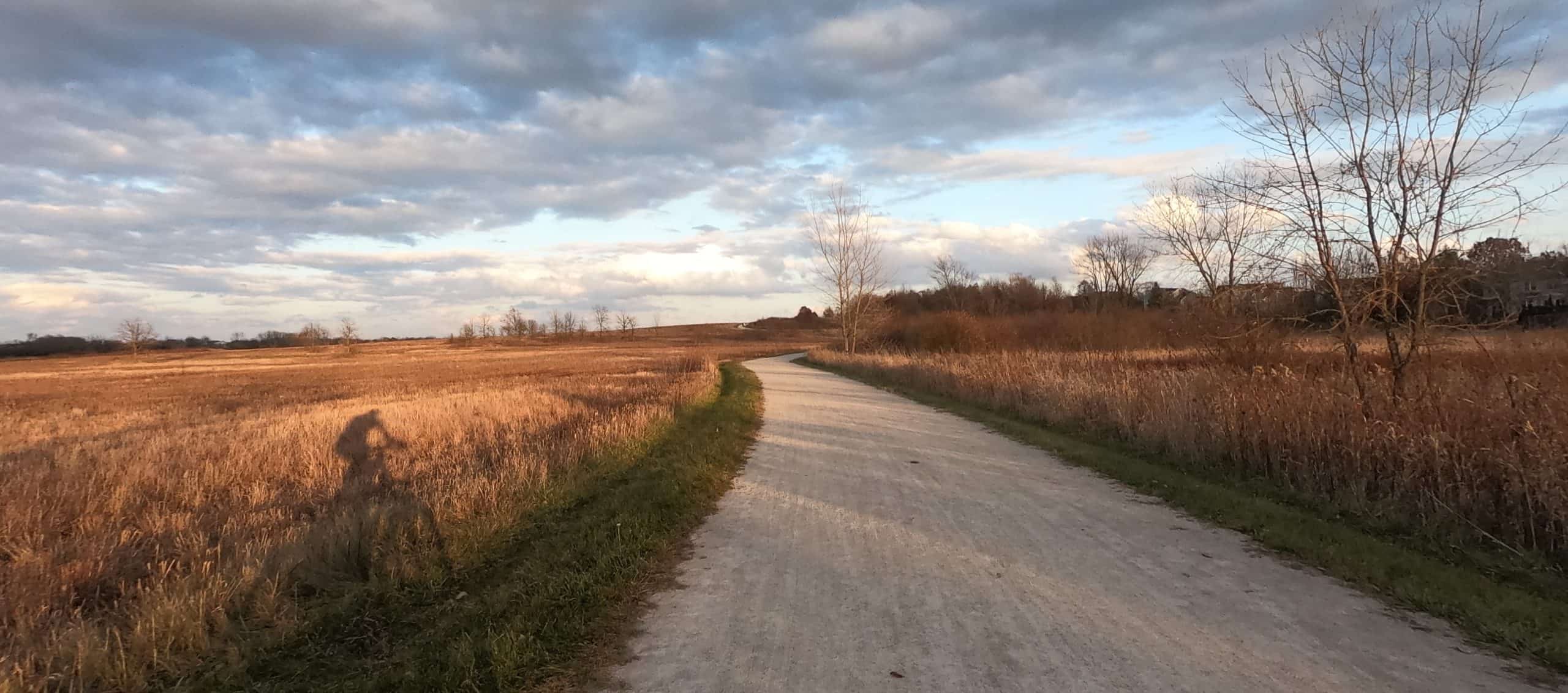 Looking down a gravel trail amid prairie with a shadow of a cyclist on the left.