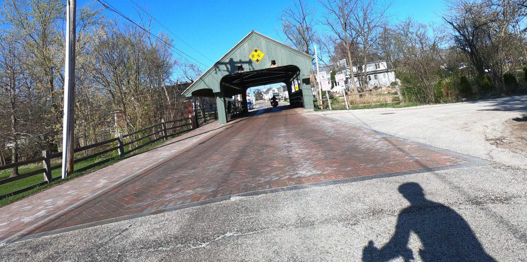 Looking ahead at a road with some brick pavement leading to a covered bridge ahead, on a sunny day.