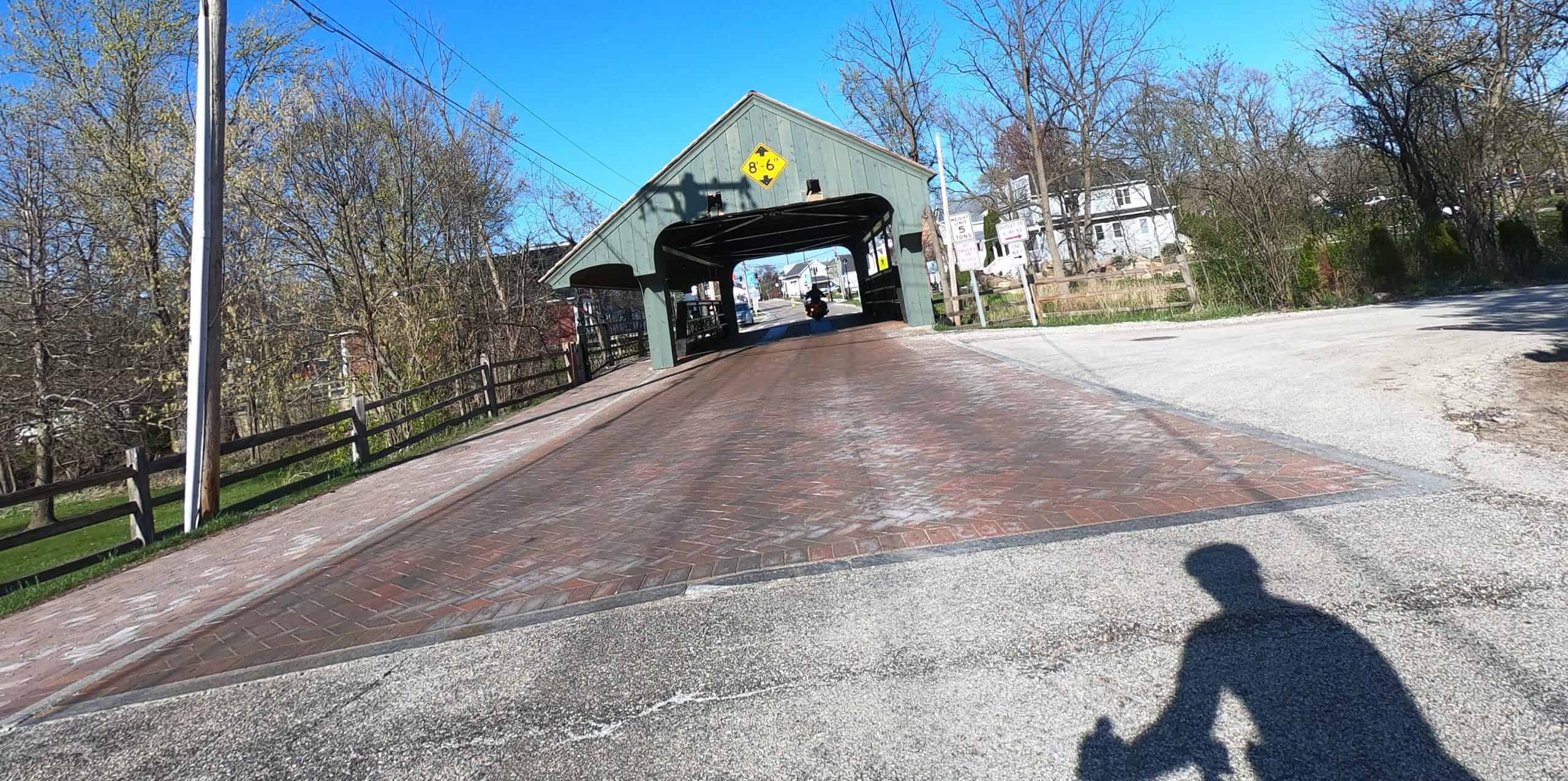Looking ahead at a road with some brick pavement leading to a covered bridge ahead, on a sunny day.