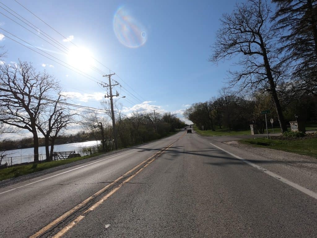 Looking down a two-lane road with water seen on the left, short trees seen on either side down the road, the sun at the top left and blue skies.