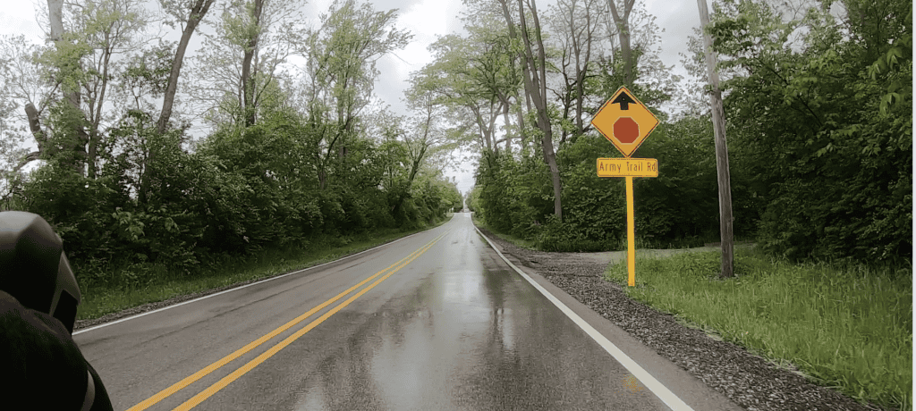 Looking ahead on a wet, two-lane road lined with trees, with a yellow traffic sign coming on the right.