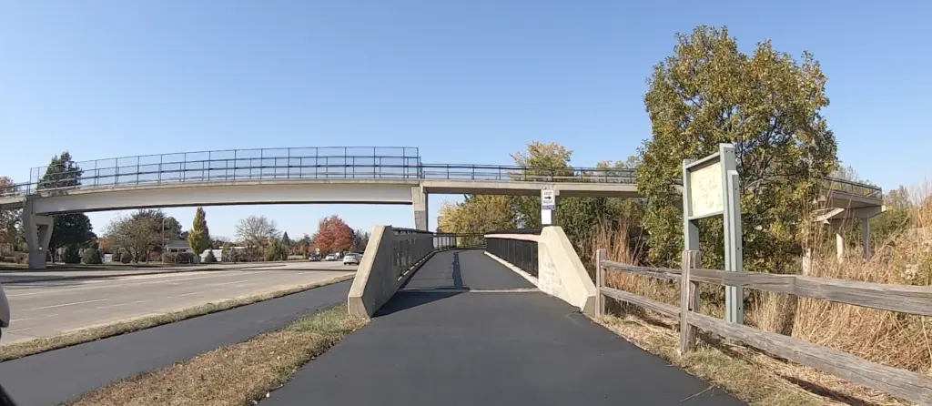 Looking down a trail leading to a bicycle-pedestrian bridge over a busy four-lane street on the left.