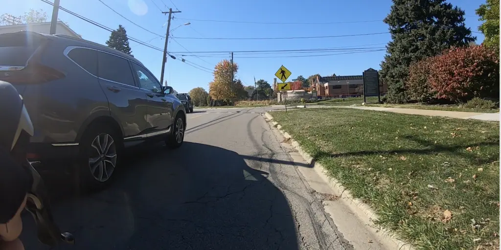 An SUV rolls just on the left as we look down a street with lawn, bushes and trees on the right and a pedestrian crosswalk ahead.