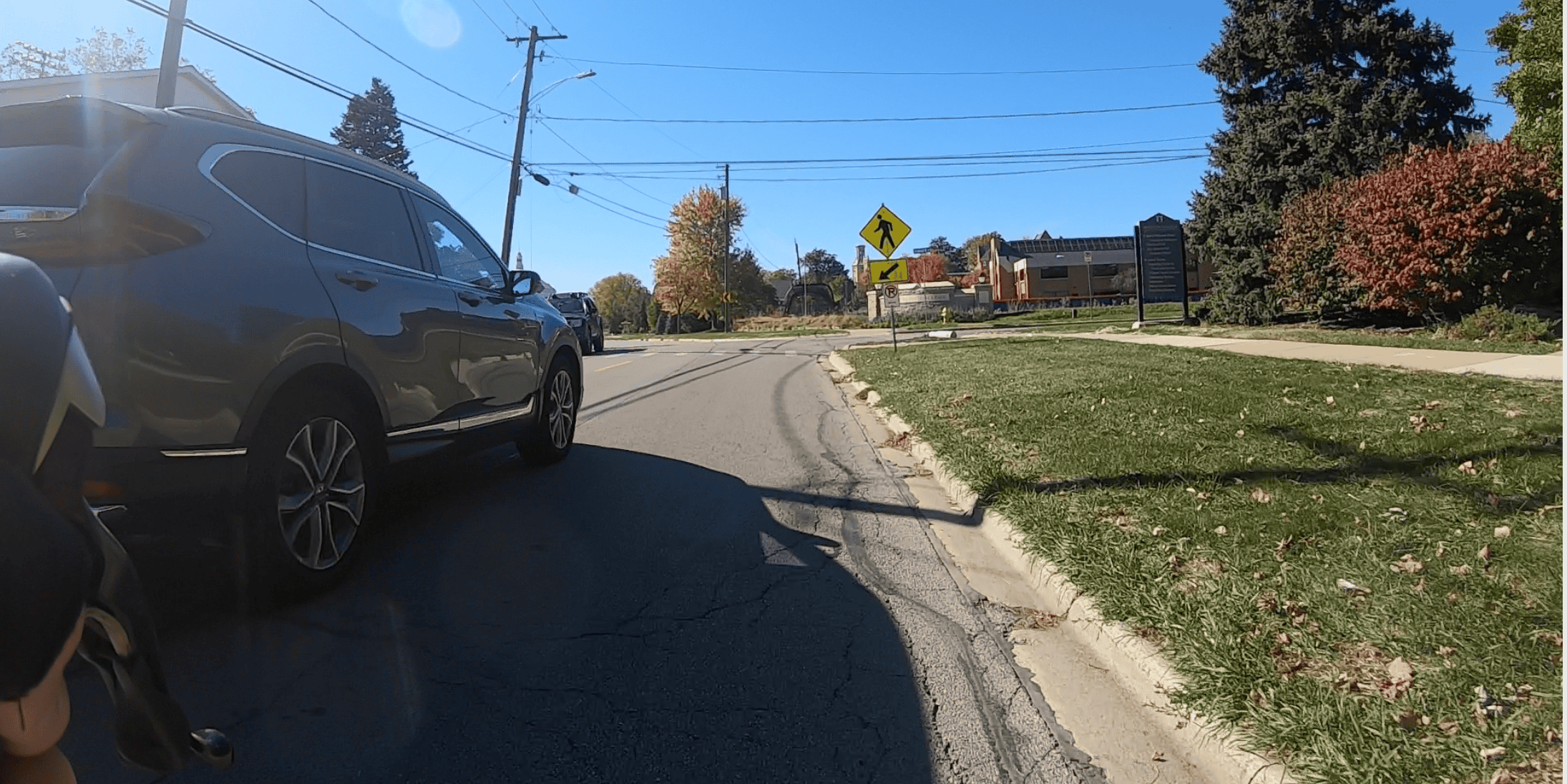 An SUV rolls just on the left as we look down a street with lawn, bushes and trees on the right and a pedestrian crosswalk ahead.
