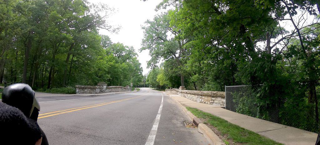 Looking down a two-lane paved road with a shoulder, on a bridge over a ravine, tall green trees in the background.