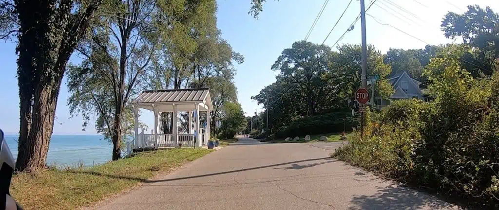A view down a road with a tall tree, a shelter and a big blue lake on the left, with an intersection and trees ahead on the right.