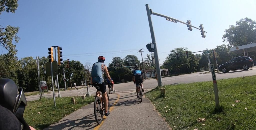 Bicyclists in cycling outfits on a paved trail come up on a busy intersection.