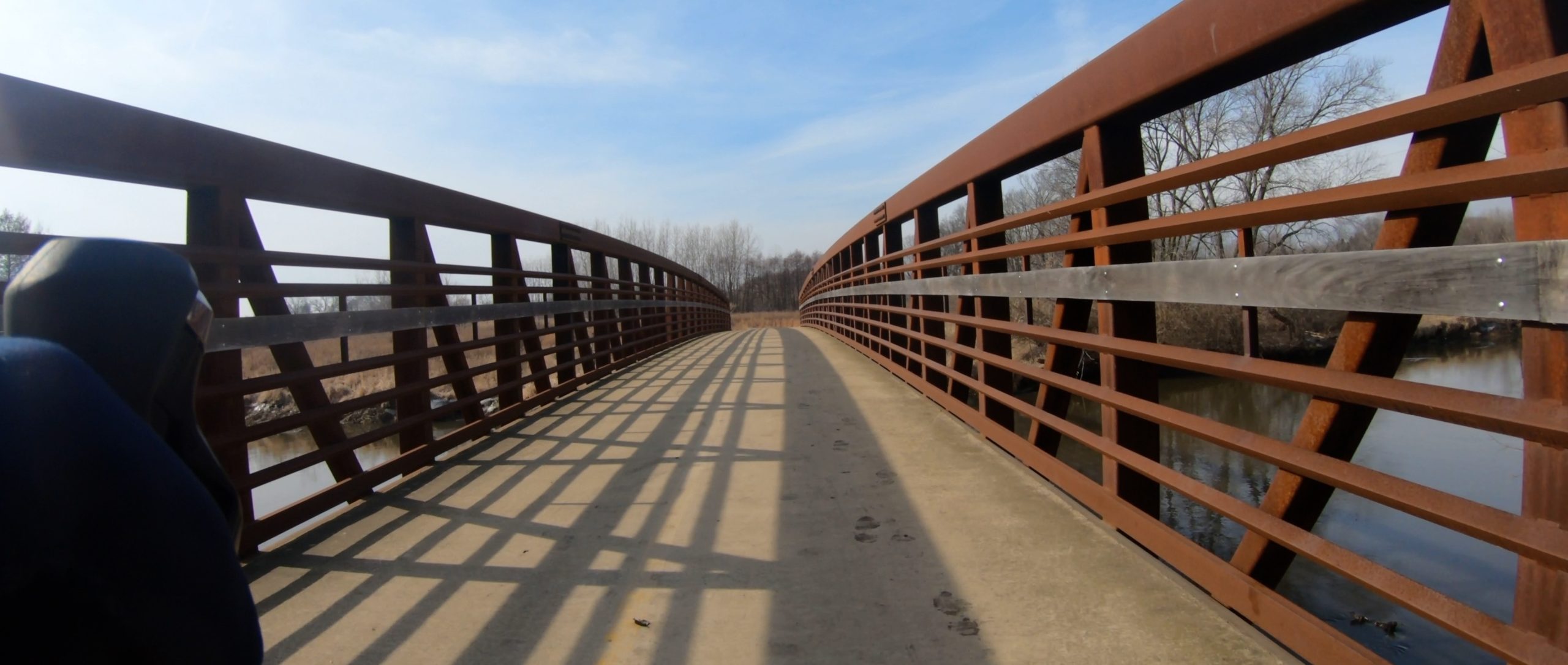Looking down a cement path on a bridge with burnt-orange fencing on a sunny day with prairie and trees with no leaves in the background.