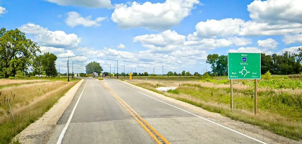 Looking down a two-lane paved road with a sign on the right indicating a roundabout ahead, and prairie and grass on the sides.