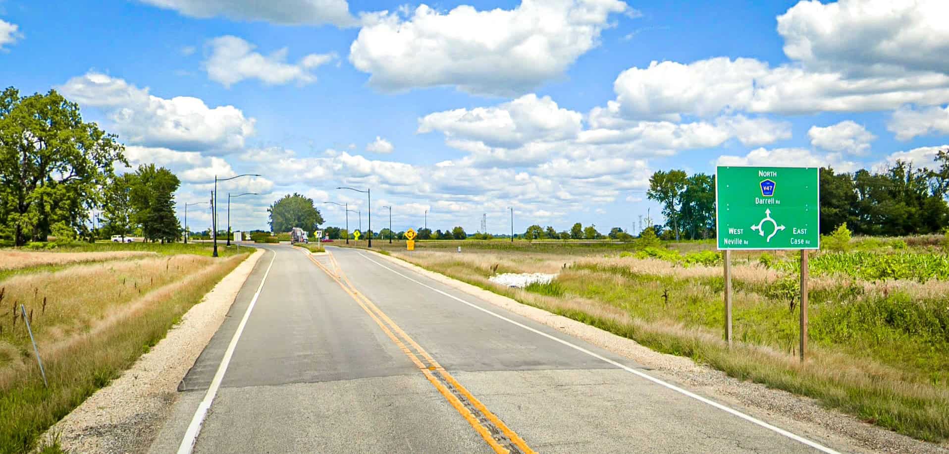 Looking down a two-lane paved road with a sign on the right indicating a roundabout ahead, and prairie and grass on the sides.