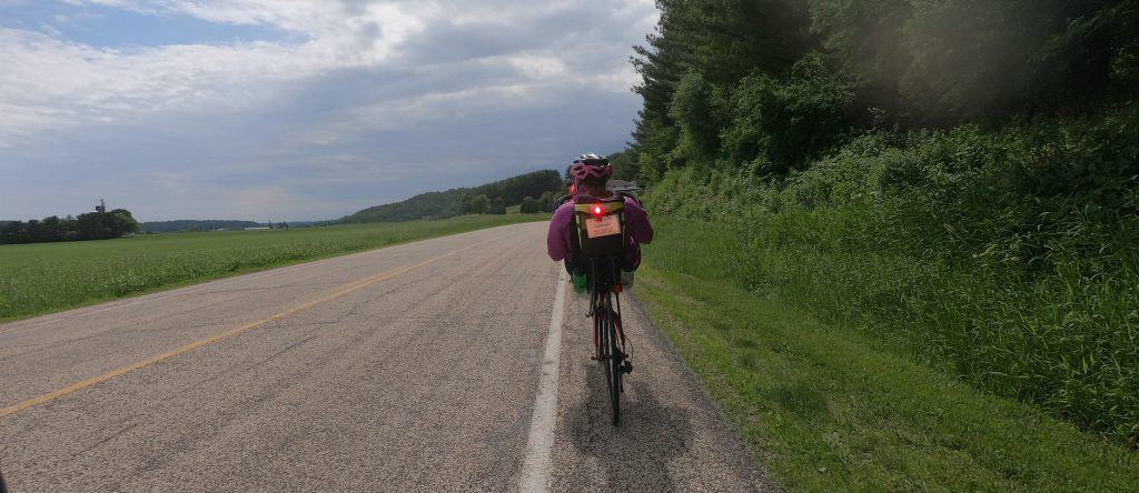 A recumbent bicyclist rides on a shoulder of a paved two-lane road beside green bushes and trees with a green field of crops on the left.