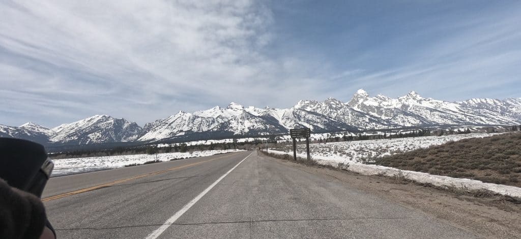 Looking down a paved road with snow-capped mountains far in the background, snow-covered prairie on the sides.