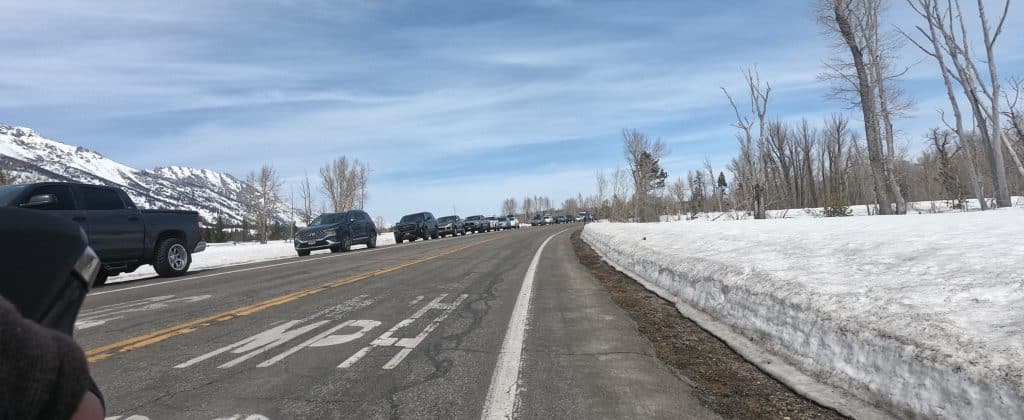 Looking down a paved road amid snow-covered open land, with a line of cars seen parked to the left.