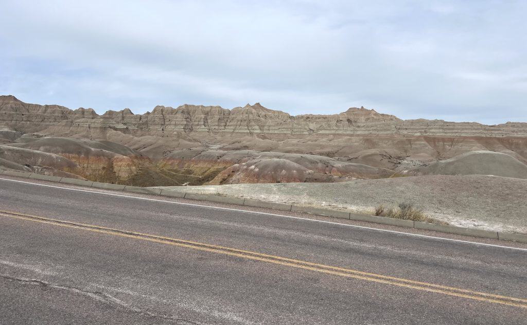 Looking at some of the Badlands National Park across a two-lane paved road under a cloudy sky.