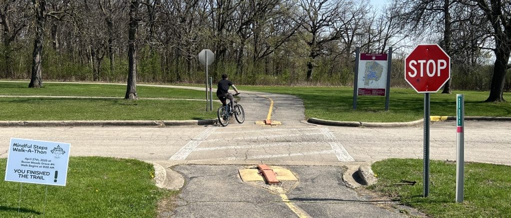 A bike rider is seen entering a striped paved bike trail around a folded-down bollard from a parking lot road in a forest preserve.
