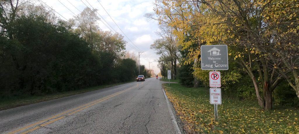 Looking down a 2-lane tree-lined road with a "Welcome to Long Grove" sign, and a car and bicyclist down the road.