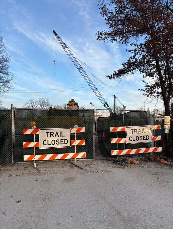 Two "Trail Closed" signs on orange construction barricades stand in front of a nearly solid dark green fence, with construction equipment behind it and tall trees on the sides.