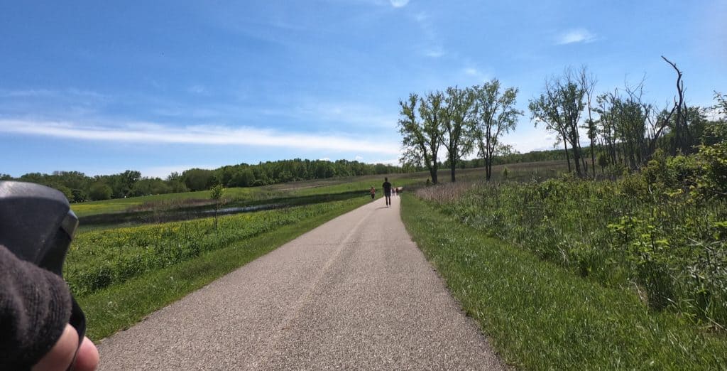 Looking down a paved trail bisecting open green prairie and wetlands under a blue sky, with a skater and walkers far ahead, a few trees in the background, a forest in the far background.