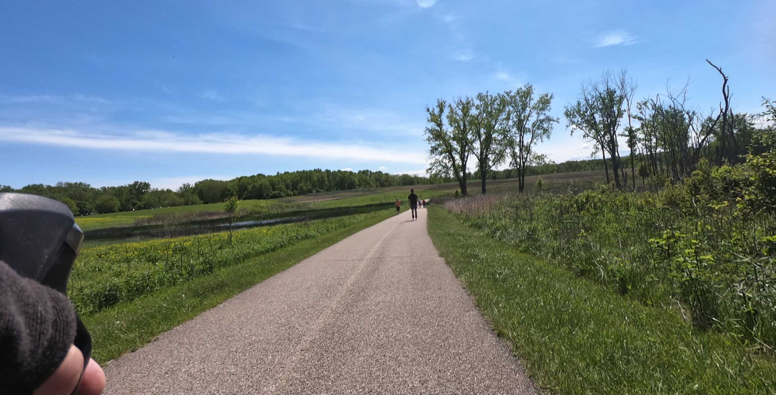 Looking down a paved trail bisecting open green prairie and wetlands under a blue sky, with a skater and walkers far ahead, a few trees in the background, a forest in the far background.
