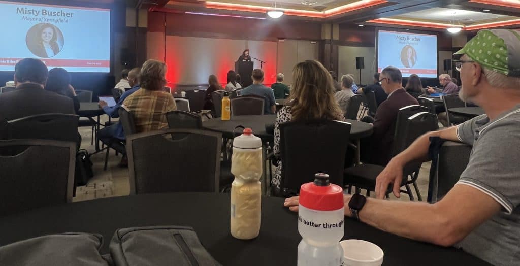 A woman is giving a presentation at a conference, in a dimly lit room full of people at round tables.