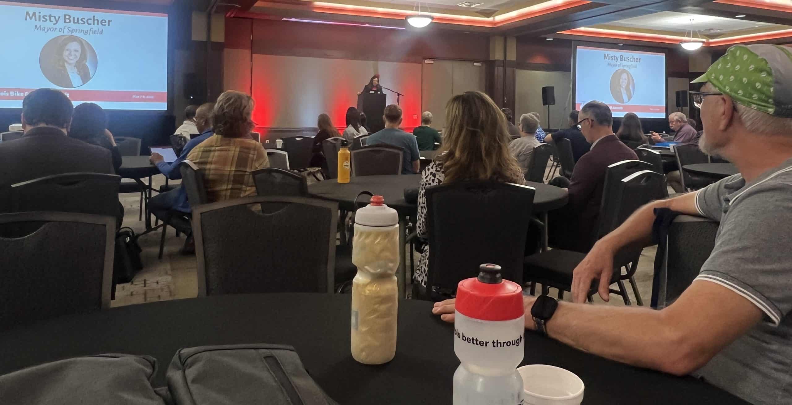 A woman is giving a presentation at a conference, in a dimly lit room full of people at round tables.